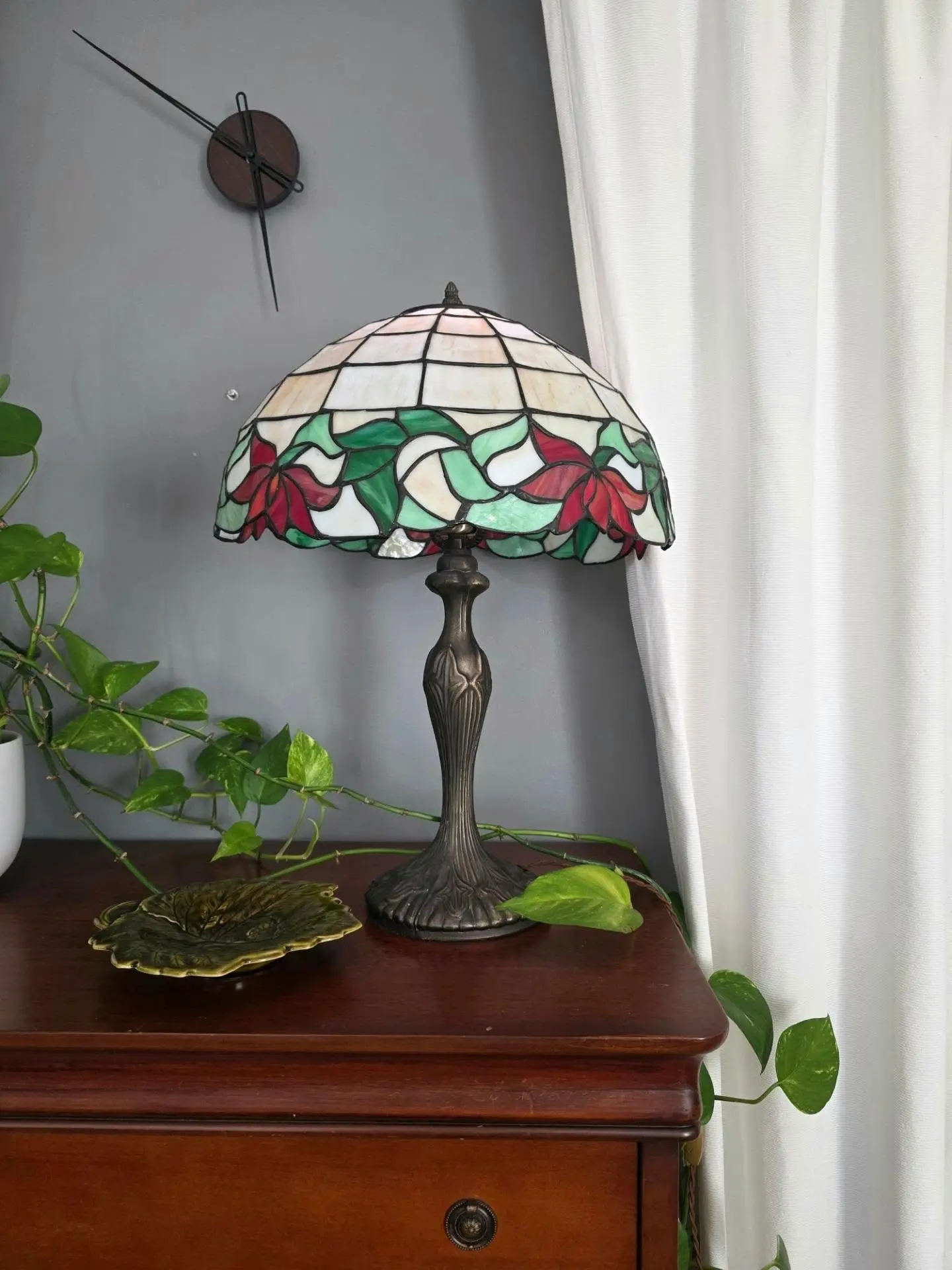 Lampe à abat-jour en vitrail fleuri, rouge et vert, sur une commode en acajou, avec un pothos et un vide-poche en forme de feuille.